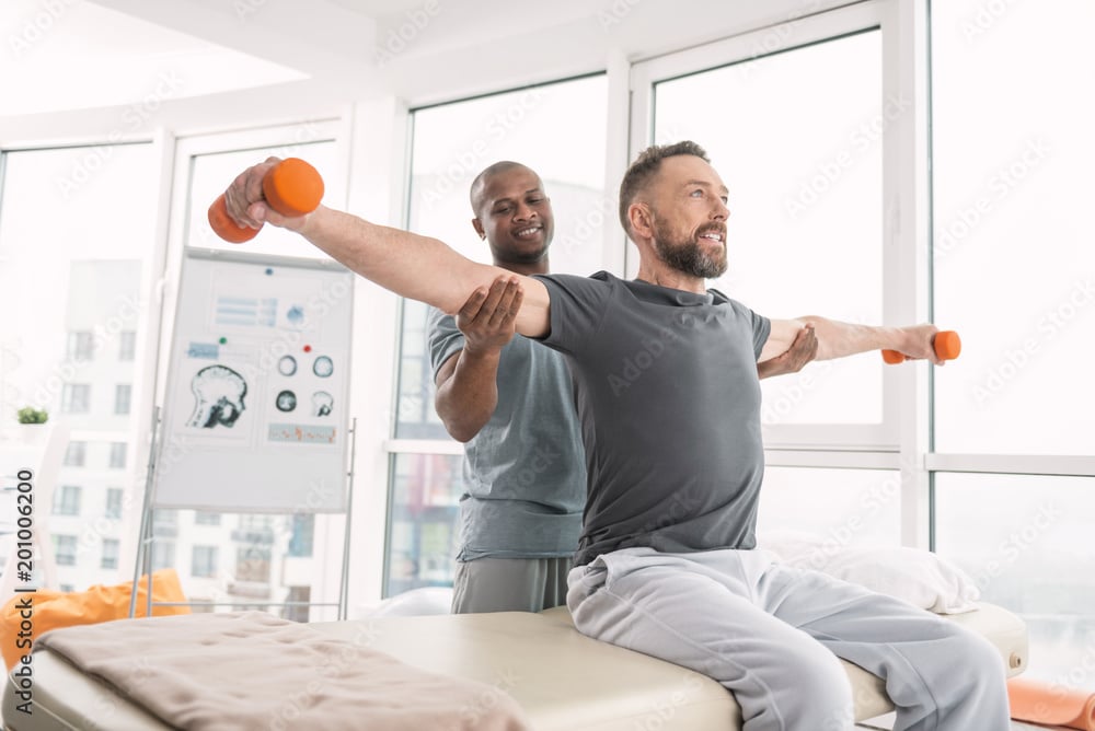 Professional supervision. Nice positive doctor standing behind his patient while fixing his hands in the right position at Well in Montclair for Emsella in Montclair, NJ.