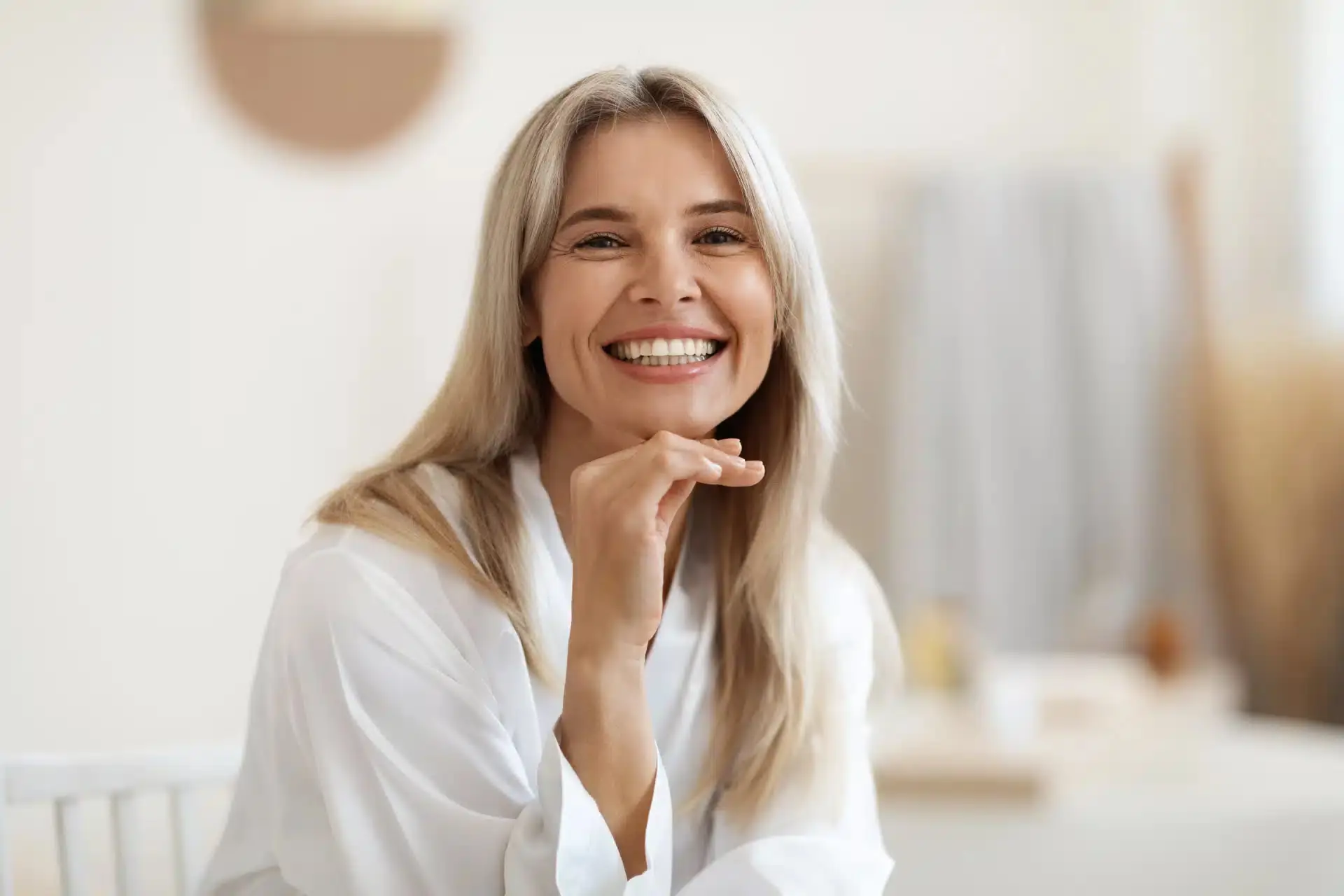 Smiling middle-aged woman with long blonde hair wearing a white robe, sitting indoors in a bright, softly lit room.