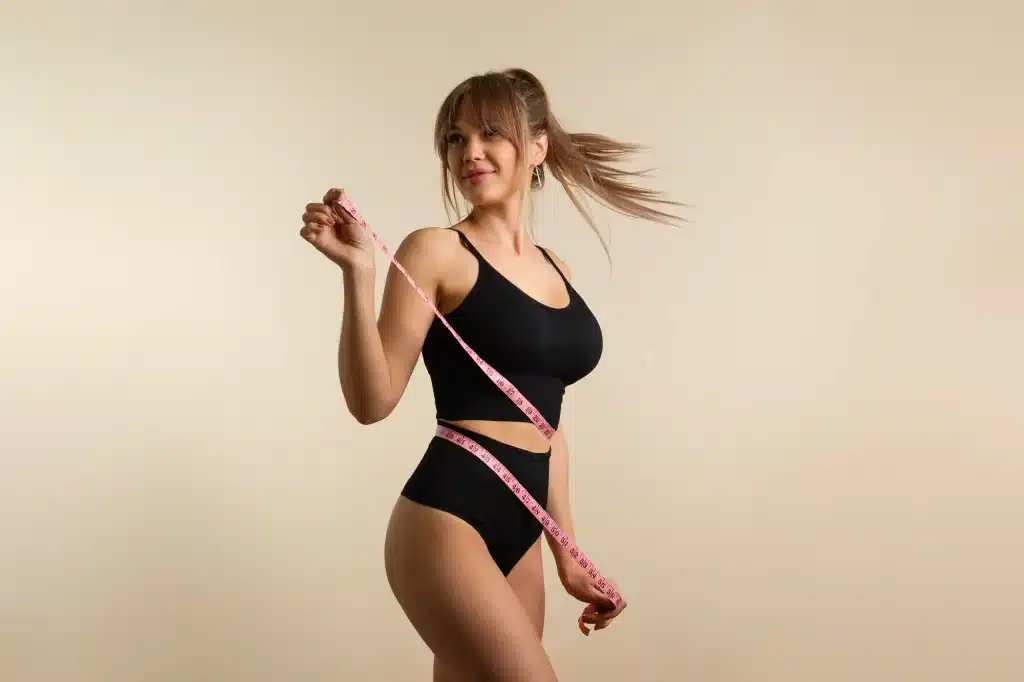 Fit woman in black activewear smiling while holding a pink measuring tape around her waist, against a beige background. representing successful weight loss with Tirzepatide at Well in Montclair in Montclair NJ.