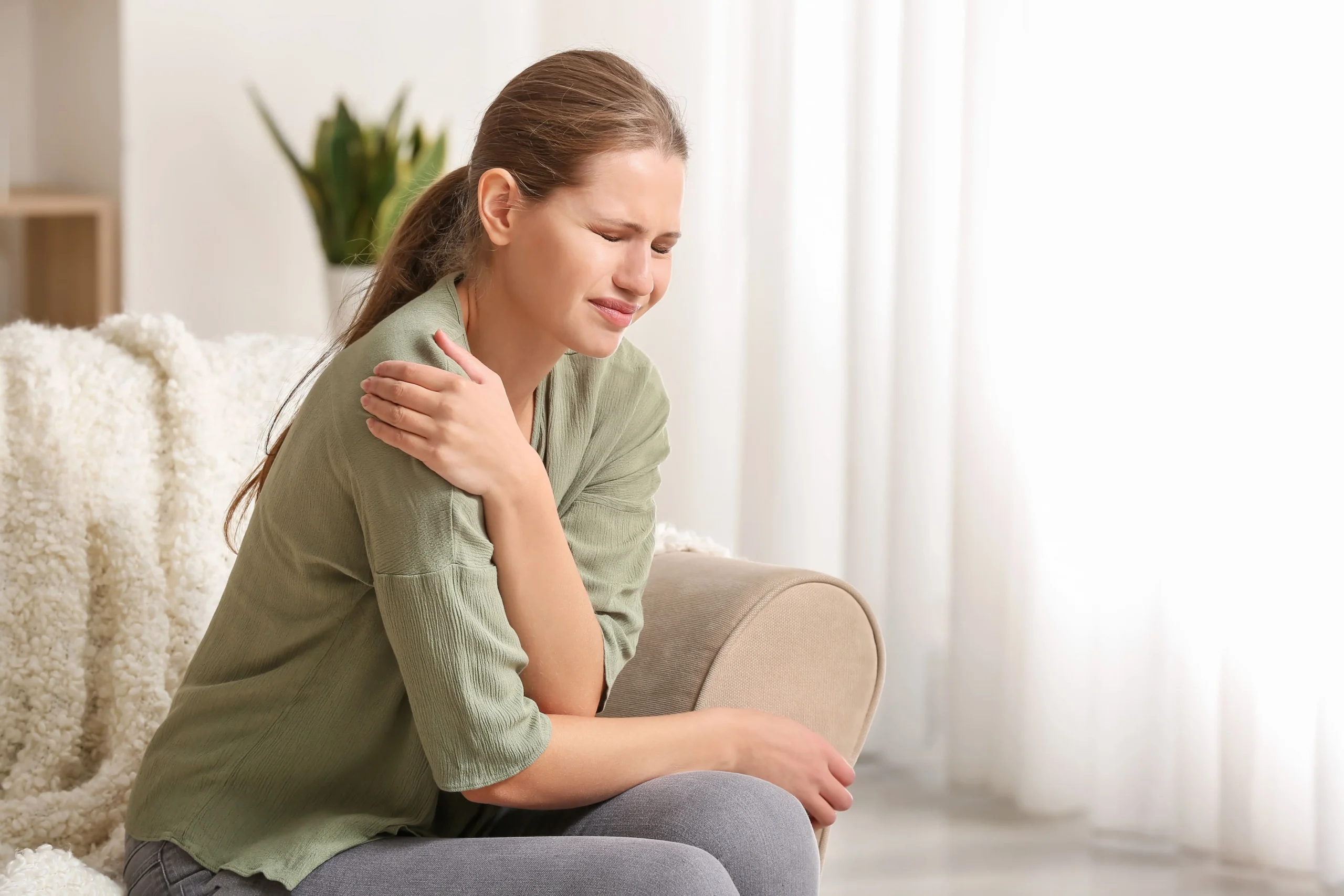 A woman sitting on a living room sofa clutching an aching shoulder in discomfort, illustrating non-invasive recovery with shockwave therapy for shoulder pain in Montclair, NJ.