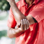 Close-up of a person in a coral polo shirt clutching a sore arm, highlighting pain relief through shockwave therapy for tennis elbow in Montclair, NJ.