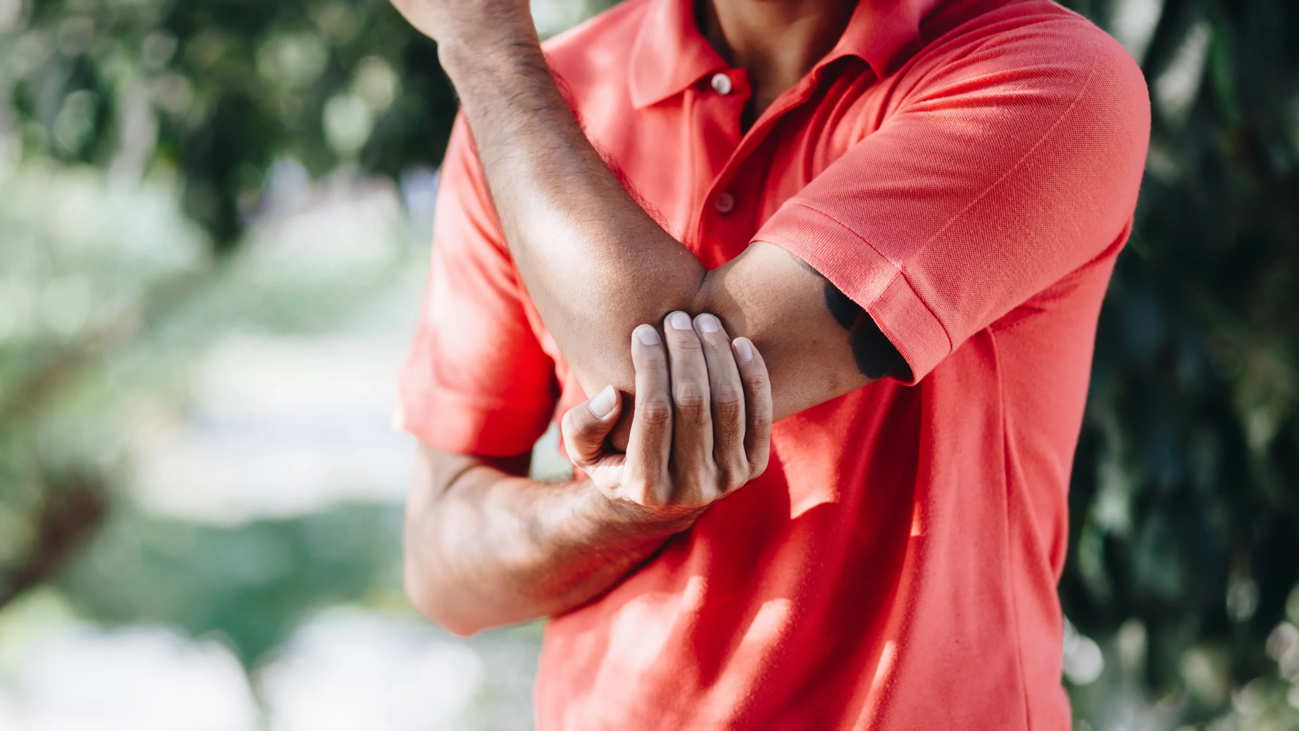 Close-up of a person in a coral polo shirt clutching a sore arm, highlighting pain relief through shockwave therapy for tennis elbow in Montclair, NJ.