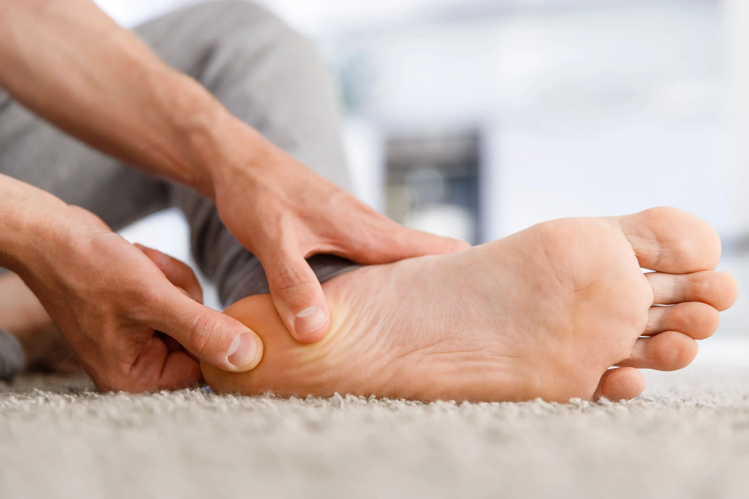 A person seated on a carpeted floor pressing thumbs into the heel and arch of a bare foot to relieve discomfort, illustrating treatment using shockwave therapy for plantar fasciitis in Montclair, NJ.